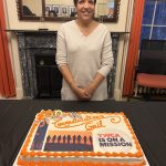 Gail Fortes smiling infront of a white cake with orange frosting on the edges, an image of her, a siloutte of women standing in front of an orange sunset, the YWCA is on a mission logo, and text reading "Congratulations Gail"