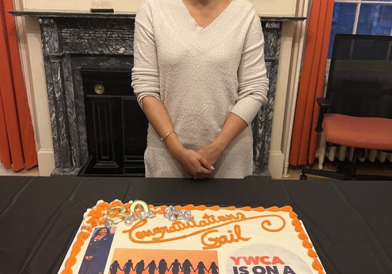 Gail Fortes smiling infront of a white cake with orange frosting on the edges, an image of her, a siloutte of women standing in front of an orange sunset, the YWCA is on a mission logo, and text reading "Congratulations Gail"