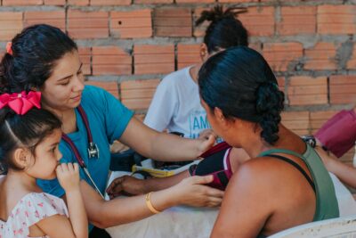 A female healthcare worker wearing teal scrubs and a stethoscope wraps a maroon blood pressure cuff around a woman’s arm. A young girl with a large pink bow in her hair sits nearby, looking on, while another person is seated in the background against an unfinished brick wall.